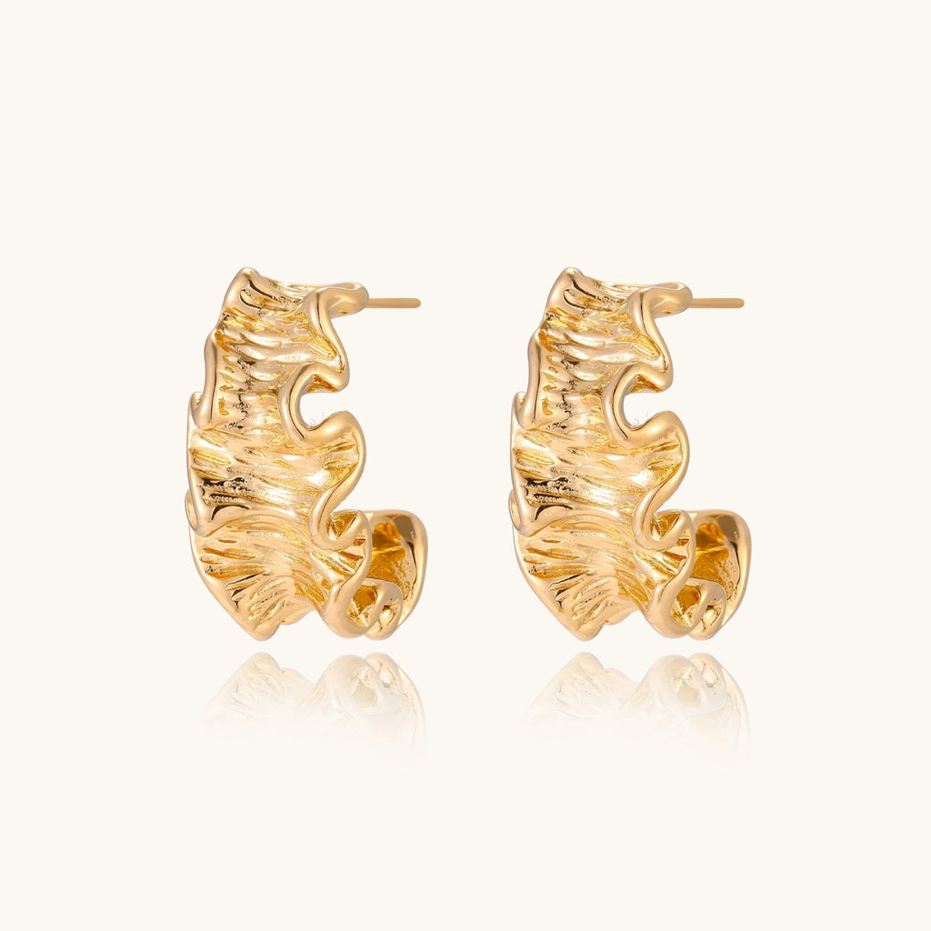 Pair of gold huggie hoops with sculpted wave texture and dimensional ripples on neutral background.