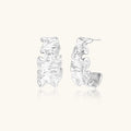 Textured white gold-plated hoop earrings with flowing wave pattern against cream background, showing dimensional rippled surface.