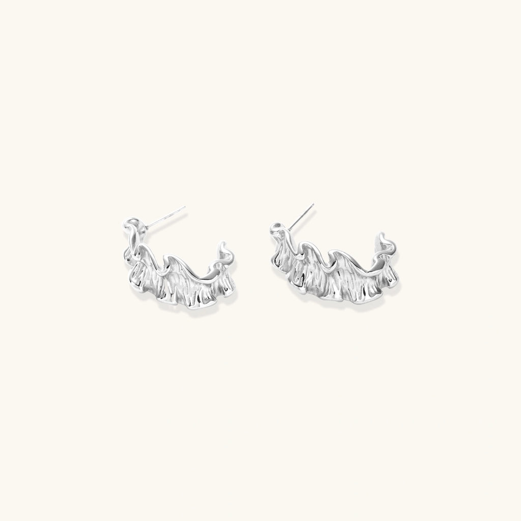 Textured white gold-plated hoop earrings with flowing wave pattern against cream background, showing dimensional rippled surface.
