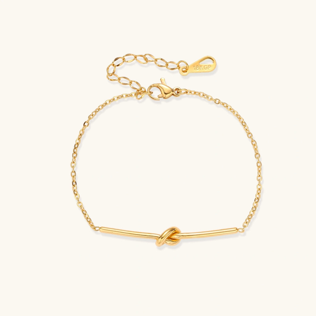 Delicate gold chain bracelet with minimalist bar and centered knot detail on cream background